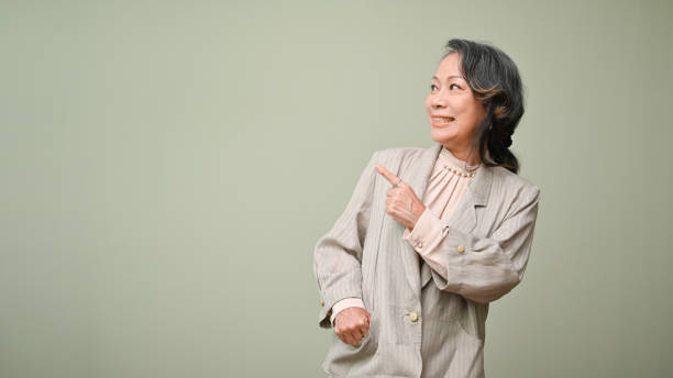 Beautiful 60s senior asian aged woman pointing index finger, smiling and looking aside stock photo