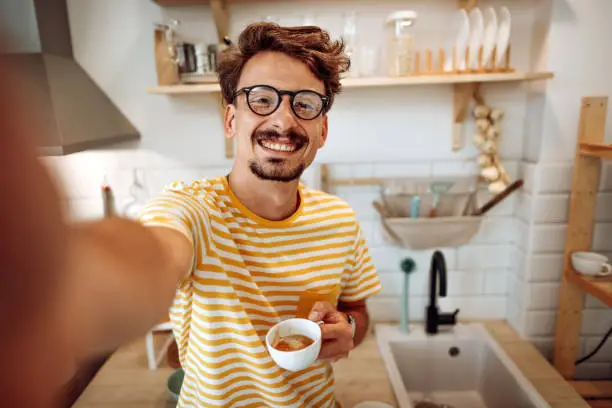 Young casually clothed man taking a selfie in his kitchen with a cup of coffee