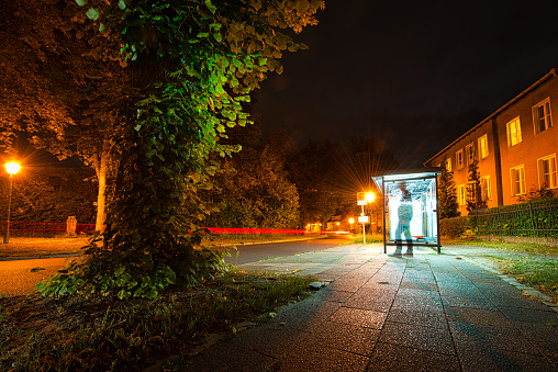This is a long-term night photograph of a bus stop. A passing car is only displayed as a light track. A man is waiting for the bus, but is only shadowy. The street lighting this summer evening with the reddish light forms a beautiful contrast to the green of the plants.