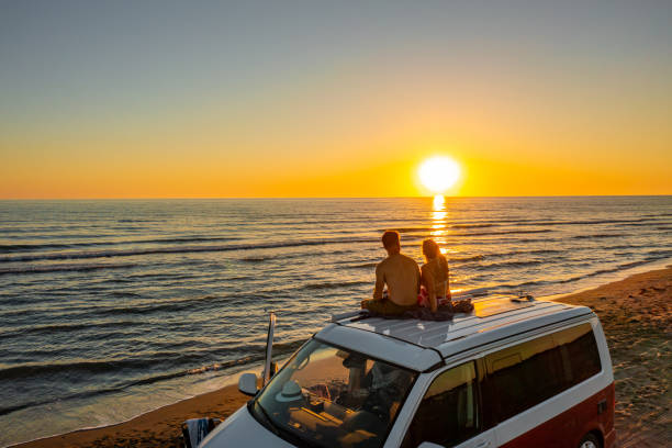 couple watching sunset while sitting on roof of campervan at beach - fordonsboende bildbanksfoton och bilder