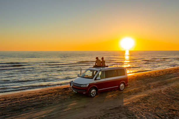 Couple sitting on roof of campervan at beach while enjoying sunset stock photo