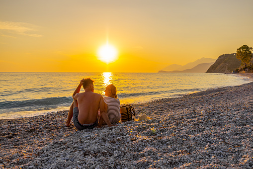 Rear view of couple relaxing at beach while enjoying sunset during summer vacation