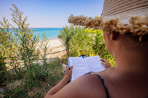 Close-up of tourist wearing hat reading book while relaxing in garden by beach during sunny day