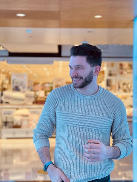 istock Man wearing a light blue sweater smiling, standing at a mall with white tiling 1432189509