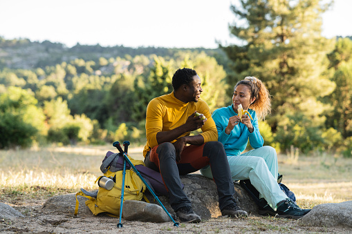 couple in nature having lunch after hiking. they are eating fruit sitting on a rock at sunset while talking.