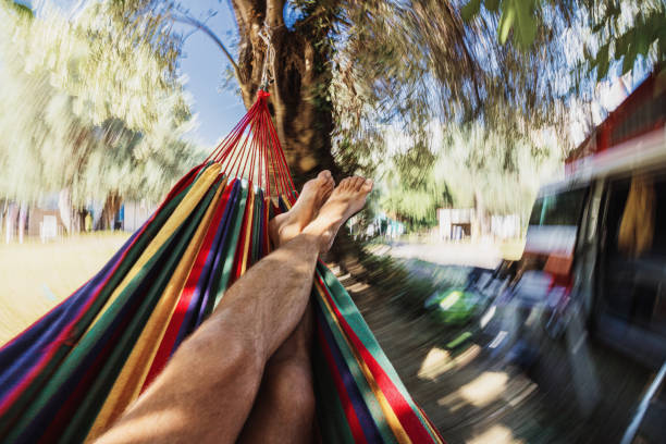 Barefoot man relaxing in hammock next to parked motor home at campsite on sunny day