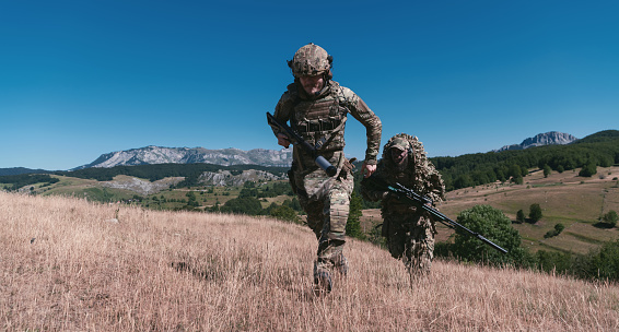 A sniper team squad of soldiers is going undercover. Sniper assistant and team leader walking and aiming in nature with yellow grass and blue sky. Tactical camouflage uniform. Hi quality stock photo.