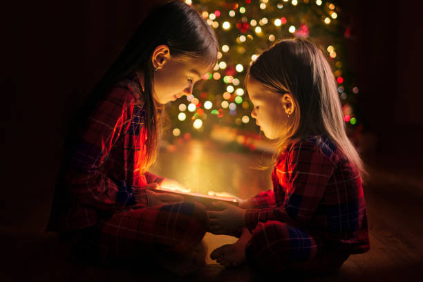 le sorelle tengono in mano una scatola con un bagliore magico dall'interno sedute sul pavimento vicino all'albero di natale. magia a natale e capodanno. un regalo di babbo natale - baby santa claus christmas sleeping foto e immagini stock