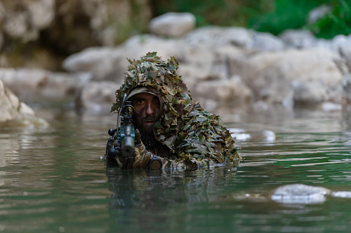 A military man or airsoft player in a camouflage suit sneaking the river and aims from a sniper rifle to the side or to target. High quality photo
