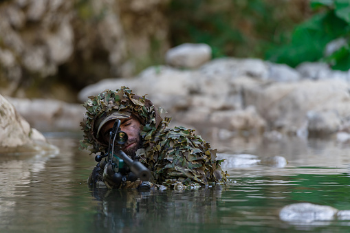 A military man or airsoft player in a camouflage suit sneaking the river and aims from a sniper rifle to the side or to target. High quality photo