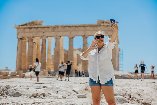 Happy influencer waving while vlogging on smart phone while exploring Parthenon temple during summer vacation