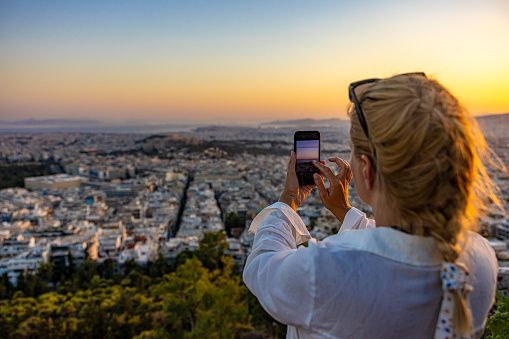 Rear view of tourist photographing cityscape through smart phone against sky during sunset