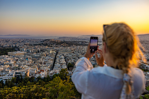 Rear view of woman photographing cityscape through smart phone against sky during sunset