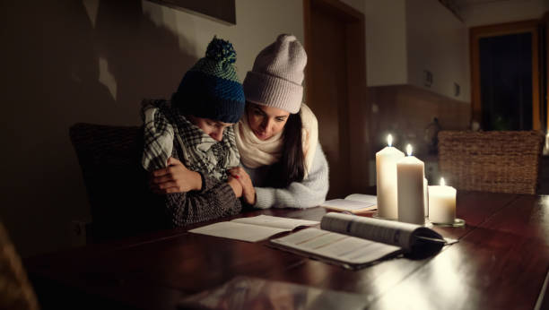 la madre tiene suo figlio tremante dal freddo studiando sotto la luce delle candele durante un balckout a causa della crisi energetica. - crisis house burning color image foto e immagini stock