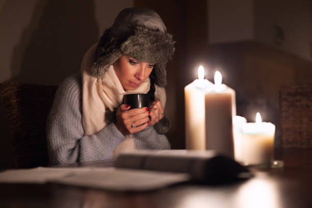 Freezing young woman in winter clothes warms her hands on cup of tea and lights with candles as energy blackouts cause electricity outages. stock photo