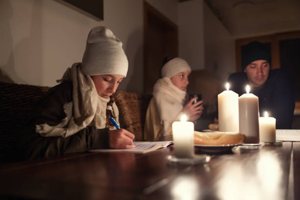Girl in winter hat and shawl studies at home at the table by candle lights while fanily experiences winter power blackout. stock photo