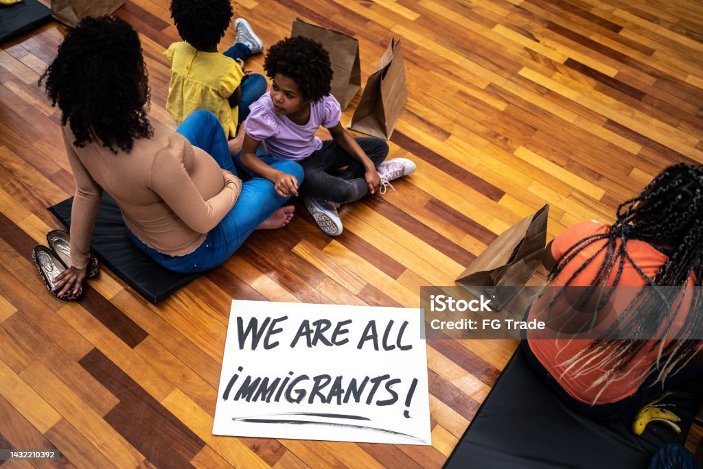 Refugee family in a sheltering sitting on the ground close to sign written "we are all immigrants!" Family Stock Photo