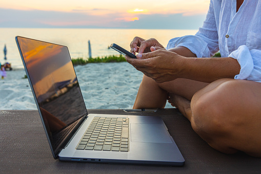 Midsection of woman using laptop and smart phone while sitting on chair at beach during sunset