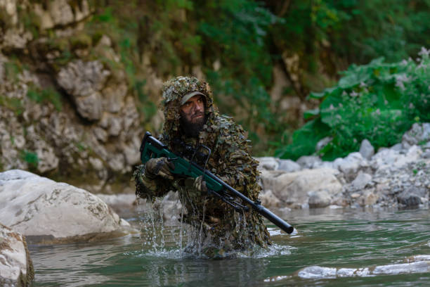 迷彩服を着た軍人またはエアガン奏者が川をこっそりと歩き回り、スナイパーライフルから横または標的を狙う。 - wet spy men suit ストックフォトと画像