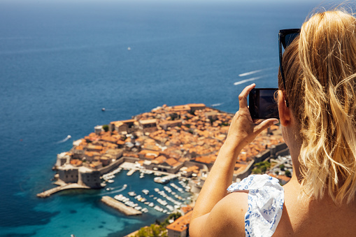 Woman taking picture of beautiful coastal town with smart phone from mountain top on sunny day