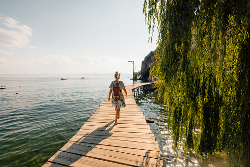 Woman with backpack walking on wooden walkway along sea on sunny summer day