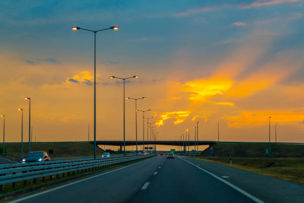 Cars driving on asphalt highway against cloudy sky during beautiful sunset