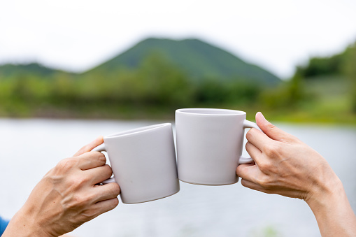 Asian man and woman friends toasting coffee cup while having breakfast and drinking coffee together in the morning. Happy family enjoy outdoor lifestyle camping in forest mountain on summer vacation.