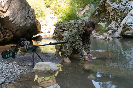 Soldier in a camouflage suit uniform drinking fresh water from the river. Military sniper rifle on the side. High quality photo