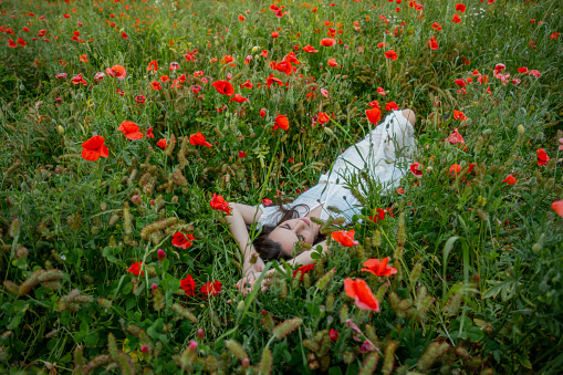 Beautiful young woman in white dress with eyes closed relaxing on poppy flowers meadow