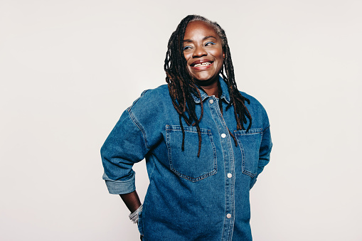 Happy woman with dreadlocks looking away with a cheerful smile while standing against a grey background. Stylish mature woman wearing a denim jacket and make-up in a studio.