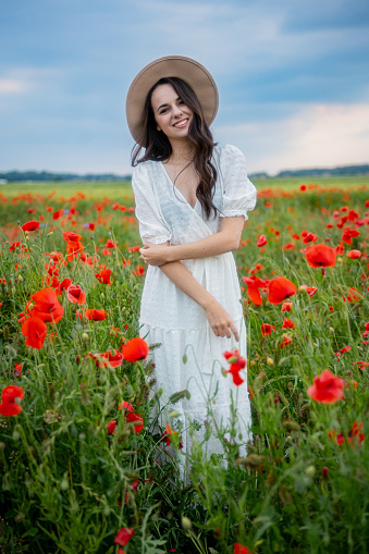 Beautiful smiling young woman in white dress and hat standing in blooming poppy field against sky