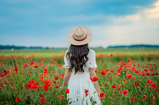 Beautiful young woman in white dress and hat standing in blooming poppy field against sky