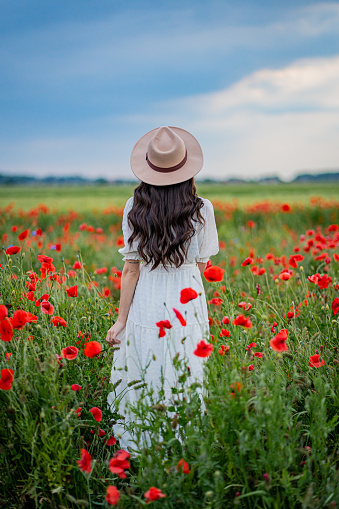 Beautiful young woman in white dress and hat standing in blooming poppy field against sky