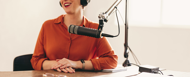 Happy woman smiling and looking away while sitting behind a microphone with a headset. Cheerful woman appearing a s a guest on a radio show. Woman co-hosting a live audio broadcast in a studio.