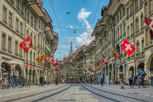 Bern, Switzerland June 03, 2019\n\nColourful Marktgasse low view on tram tracks between Swiss historical houses or buildings with fluttering flags. Cablecar or cable car railway or rail in Bern historical district