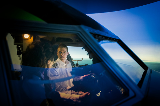 Male pilot explaining how a simulator works to a female student before a training session. Male pilot talking with woman trainee pilot sitting inside a flight simulator.