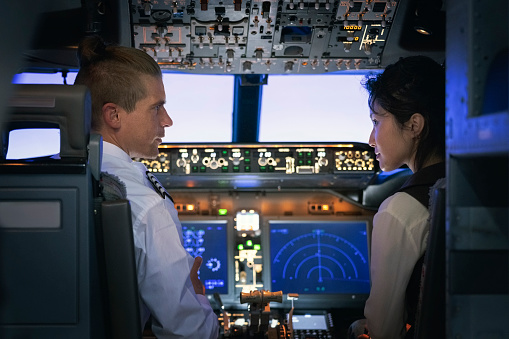 Flight instructor is explaining how a simulator works to a student before a training session inside flight simulator. Male pilot talking with woman trainee pilot sitting inside a flight simulator.