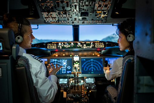 Rear view of a male pilot giving advice to female co-pilot while flying a modern jet airplane. Two pilots sitting in the cockpit and discussing route while flying an commercial airplane jet.