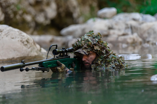 A military man or airsoft player in a camouflage suit sneaking the river and aims from a sniper rifle to the side or to target. High quality photo
