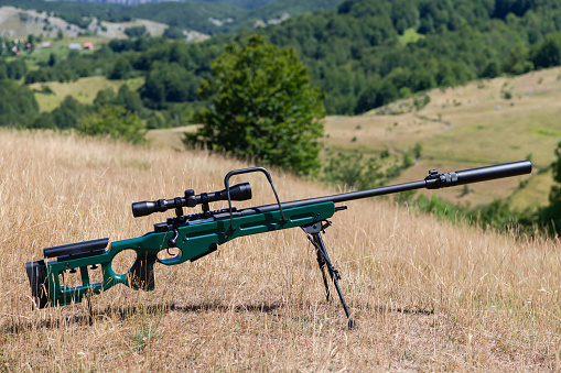 A green military sniper rifle with a scope for long distance tactical modern warfare in yellow grass and blue sky in background. Hi quality stock photo.