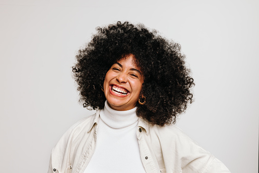 Beautiful young woman with an Afro hairstyle smiling at the camera while standing against a white background. Happy woman of color wearing her natural curly hair with pride.