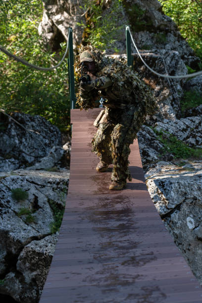 迷彩服を着た軍人またはエアガン奏者がロープブリッジをこっそりと覗き込み、スナイパーライフルから横または標的を狙う。 - wet spy men suit ストックフォトと画像