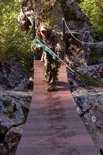 迷彩服を着た軍人またはエアガン奏者がロープブリッジをこっそりと覗き込み、スナイパーライフルから横または標的を狙う。 - wet spy men suit ストックフォトと画像