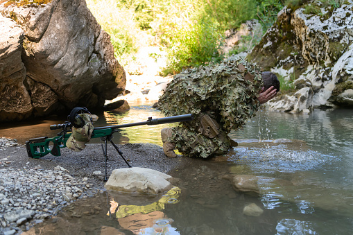 Soldier in a camouflage suit uniform drinking fresh water from the river. Military sniper rifle on the side. High quality photo