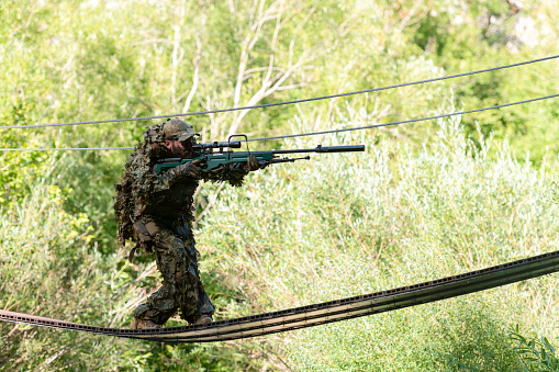 A military man or airsoft player in a camouflage suit sneaking the rope bridge and aims from a sniper rifle to the side or to target. High quality photo