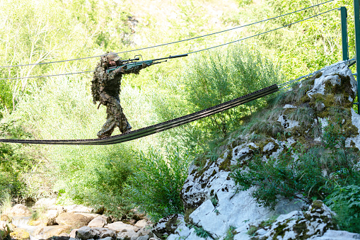 A military man or airsoft player in a camouflage suit sneaking the rope bridge and aims from a sniper rifle to the side or to target. High quality photo