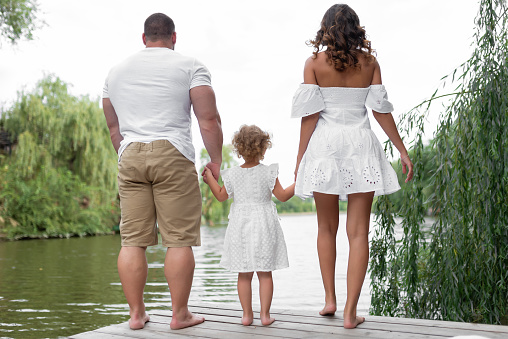 Loving family sits on a bridge by the river. Mom, dad and little daughter stand holding hands near the pond. View from the back