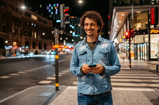 Handsome young man walking in the city street and using his smart phone at night.