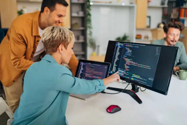 Photo of Smiling male programmer explains a coding technique to a new female colleague while sitting at a desk in the office