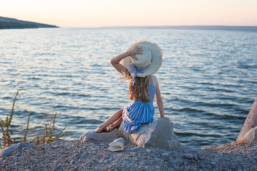 Beautiful girl with long blond hair sits on a stone on the banks of a river, lake, sea. She is looking at ocean and thinking dreamily. Girl alone outside. Girl sitting on rocks. Lonely person. Vacation on sea. Girl thinking on the beach.